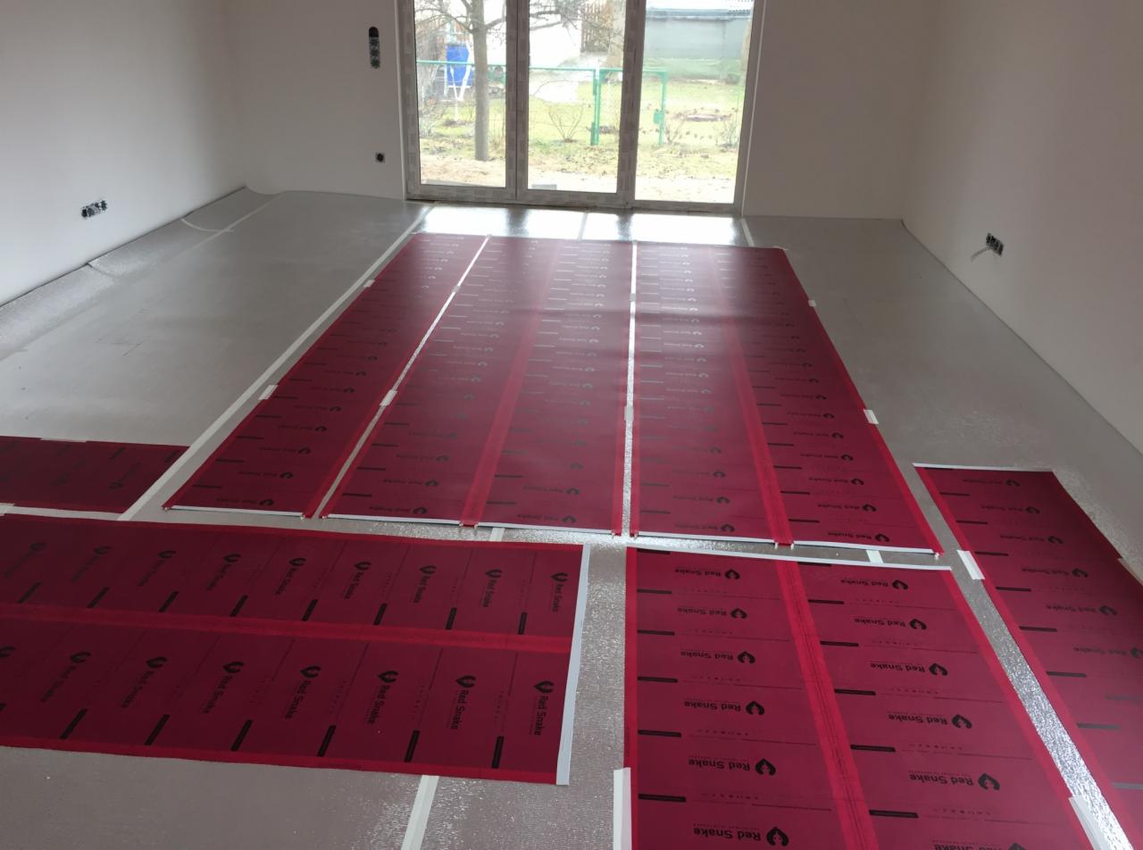 Rozłożone, czerwone panele grzewcze 'Red Snake' na podłodze z folią aluminiową, przygotowane do montażu w pomieszczeniu z oknem balkonowym.