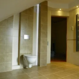 Wnętrze łazienki z beżowymi płytkami podłogowymi i ściennymi, podwieszana toaleta z chromowanym przyciskiem spłukującym, oświetlenie punktowe w suficie i wnęka z oświetleniem LED.