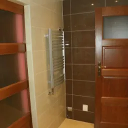 Wykończona łazienka z beżowymi i brązowymi płytkami, grzejnikiem drabinkowym i drewnianymi drzwiami z szybami.