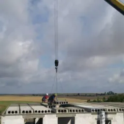 Montaż prefabrykowanych strop&oacute;w na budowie domu jednorodzinnego z widocznym dźwigiem i dwoma pracownikami w kaskach.