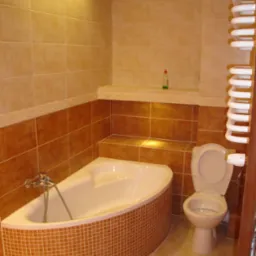 Wykończona łazienka z narożną wanną obudowaną mozaiką w odcieniach brązu i beżu, toaleta kompaktowa z otwartą klapą, grzejnik drabinkowy oraz półka z oświetleniem punktowym.