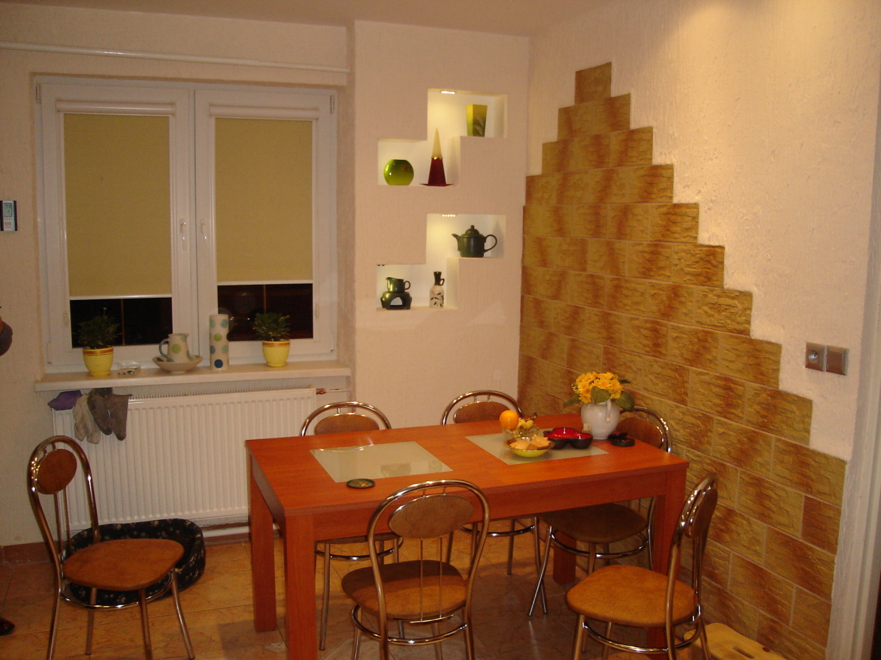 Jadalnia z drewnianym stołem i krzesłami, dekoracyjną ścianą z płytek imitujących kamień ułożonych schodkowo oraz wnękami z dekoracjami na ścianie.