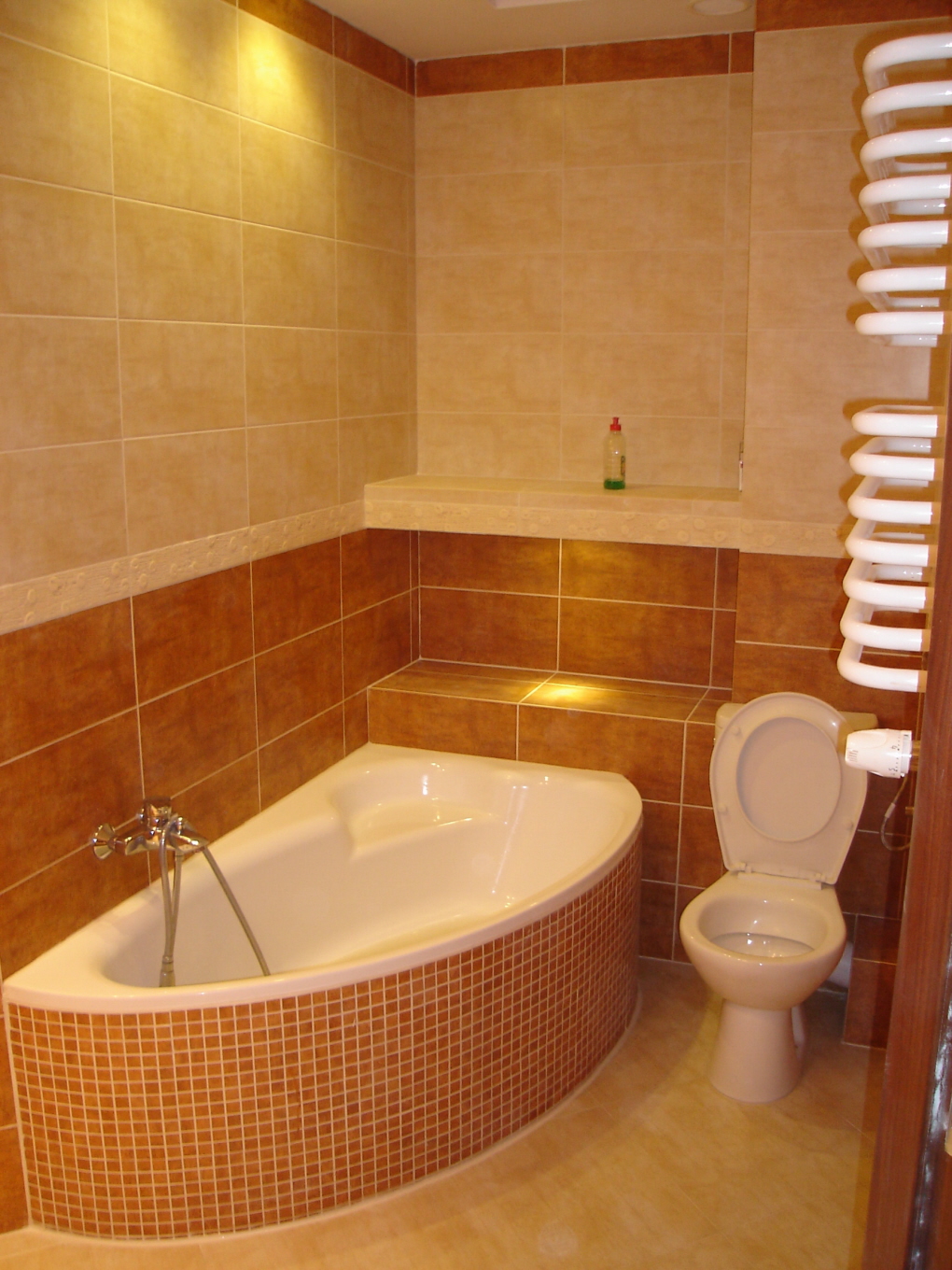 Wykończona łazienka z narożną wanną obudowaną mozaiką w odcieniach brązu i beżu, toaleta kompaktowa z otwartą klapą, grzejnik drabinkowy oraz półka z oświetleniem punktowym.