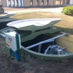 Otwarta, podziemna konstrukcja oczyszczalni z widoczną aparaturą sterującą i elementami mechanicznymi, umieszczona na trawiastym terenie z budynkiem w tle.