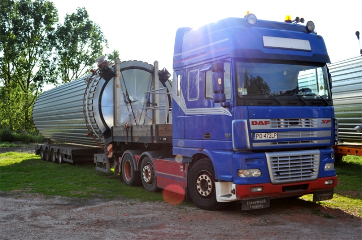 Niebieski ciągnik DAF XF z naczepą przewożącą duży, srebrny, cylindryczny zbiornik z żebrowaną powierzchnią, zaparkowany na trawiastym terenie.