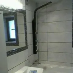 Wnętrze łazienki po remoncie: białe płytki z ciemnymi fugami, czarny panel prysznicowy z deszczownicą, umywalka z baterią i lustro z dekoracyjną ramą. Widoczne okno w tle.