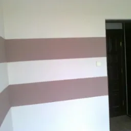 Pomieszczenie po malowaniu ścian w poziome pasy w odcieniach bieli i beżu, widoczny włącznik światła i fragment ciemnych drzwi.