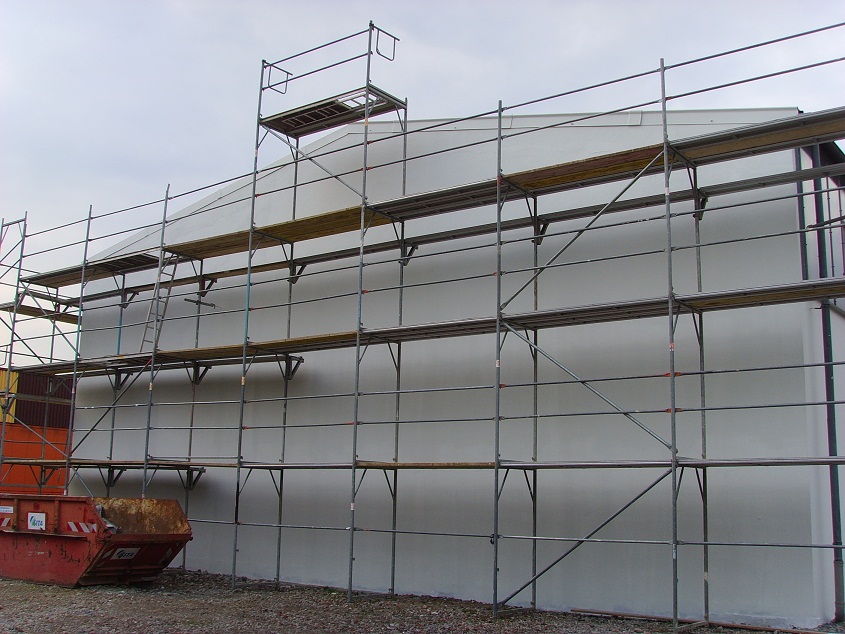 Rusztowanie metalowe wielopoziomowe ustawione przy otynkowanej ścianie budynku, z widocznym kontenerem na gruz w lewym dolnym rogu.