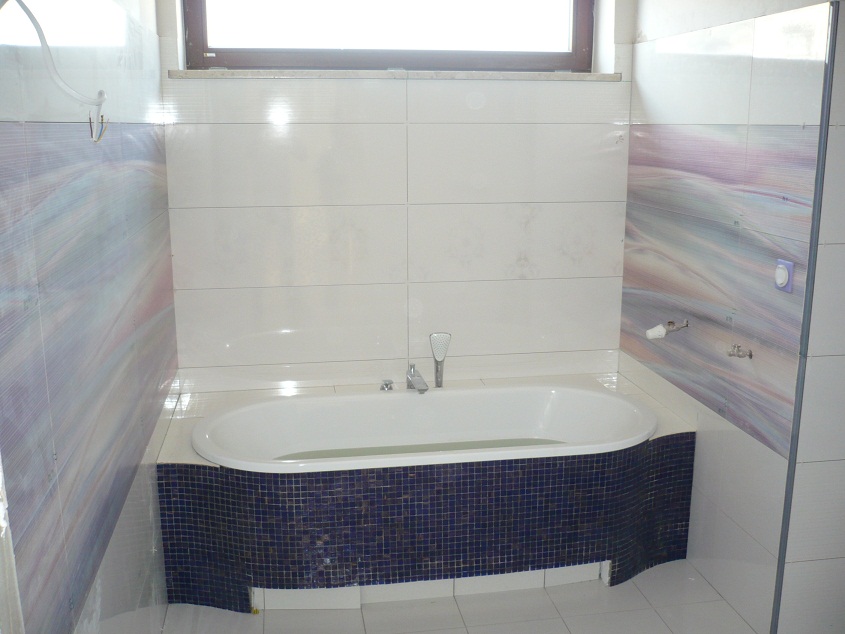 Nowoczesna łazienka z białą wanną obudowaną ciemnoniebieską mozaiką, ściany wyłożone białymi i wzorzystymi płytkami, w tle okno.