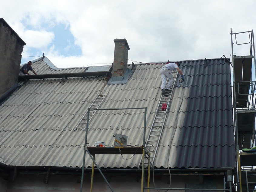 Malowanie dachu z eternitu na szaro, widoczny fragment dachu przed i po malowaniu, pracownik na drabinie, rusztowanie i komin w tle.