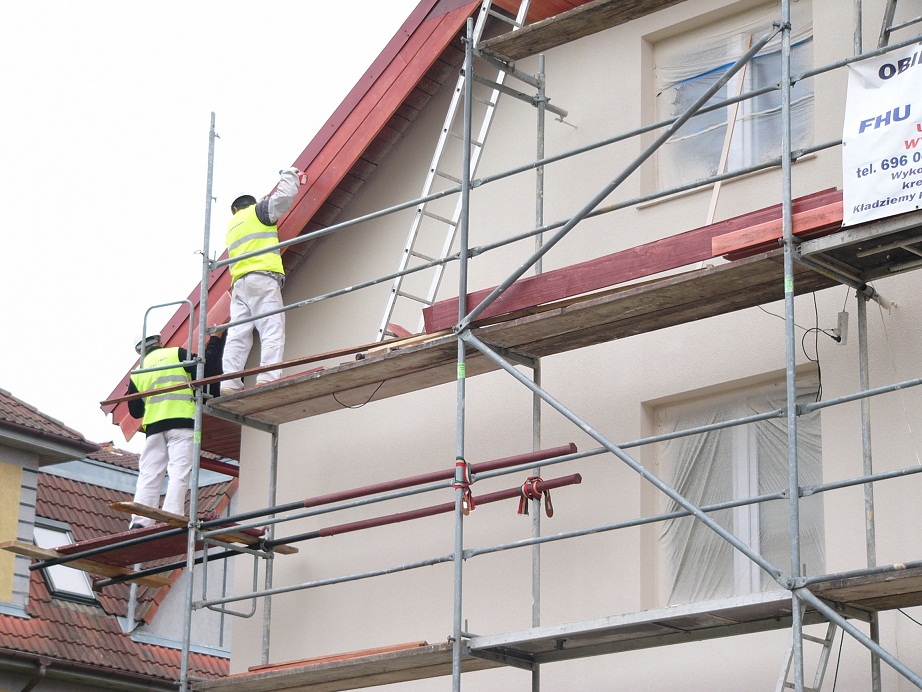 Dwóch pracowników w odblaskowych kamizelkach na rusztowaniu podczas malowania elewacji domu jednorodzinnego. Okna zabezpieczone folią malarską. Na rusztowaniu widoczny baner reklamowy firmy.