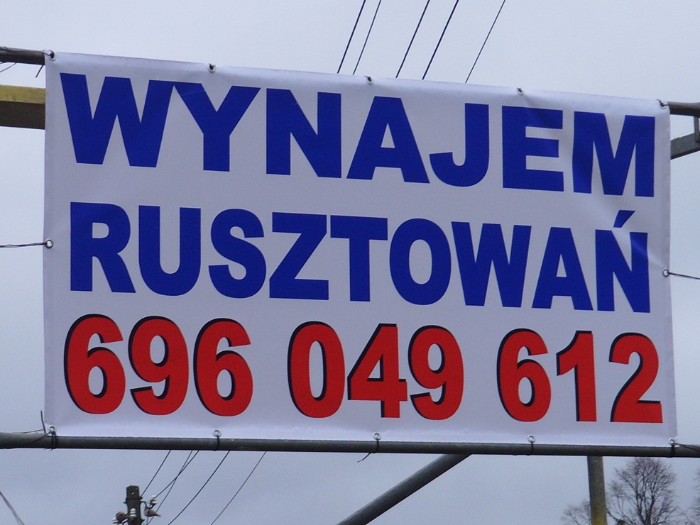 Biały baner reklamowy z niebieskim napisem 'WYNAJEM RUSZTOWAŃ' i czerwonym numerem telefonu 696 049 612, zawieszony na metalowej konstrukcji rusztowania na tle pochmurnego nieba.