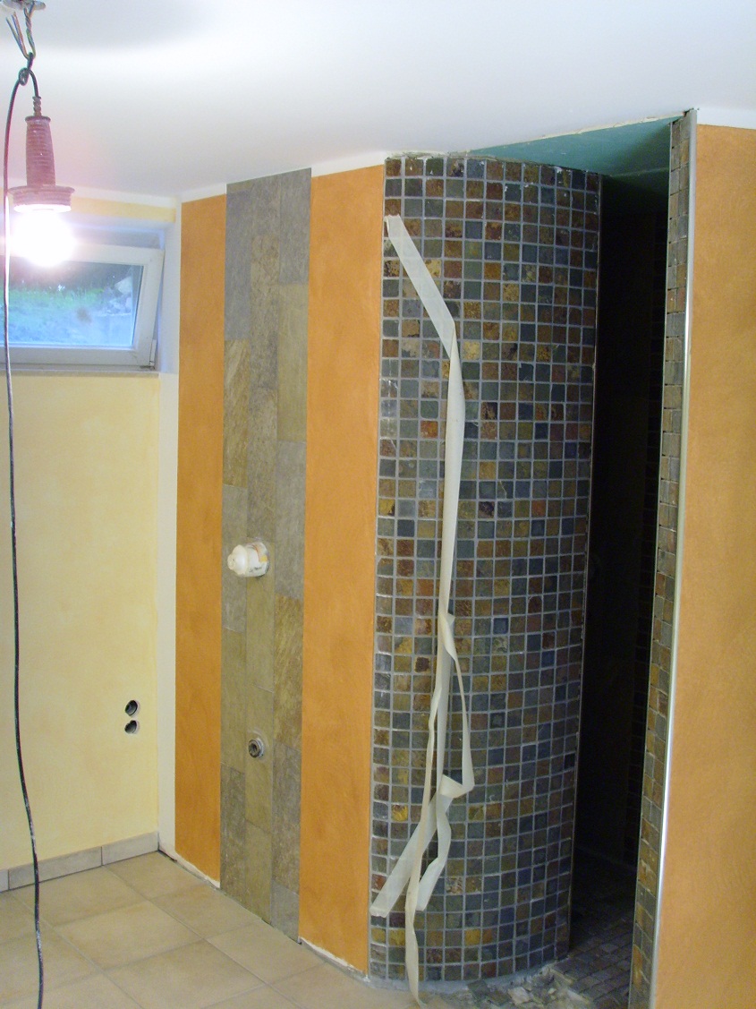 Niedokończona mozaika z małych, kolorowych płytek na zaokrąglonej ścianie kabiny prysznicowej, zabezpieczona taśmą malarską; obok fragment ściany z paskiem kamienia dekoracyjnego i żółtą farbą...
