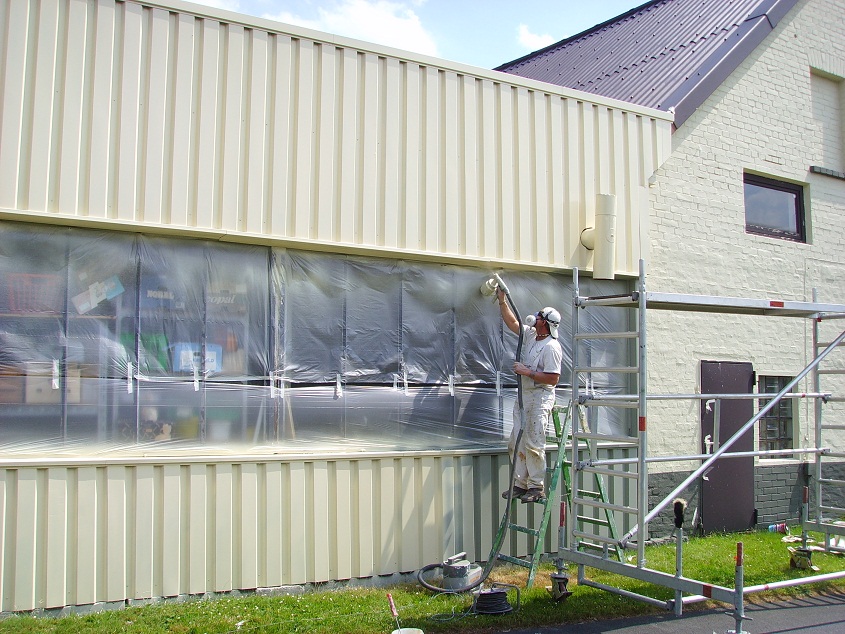 Pracownik w kombinezonie i masce maluje elewację budynku przemysłowego natryskowo z rusztowania, okna zabezpieczone folią.