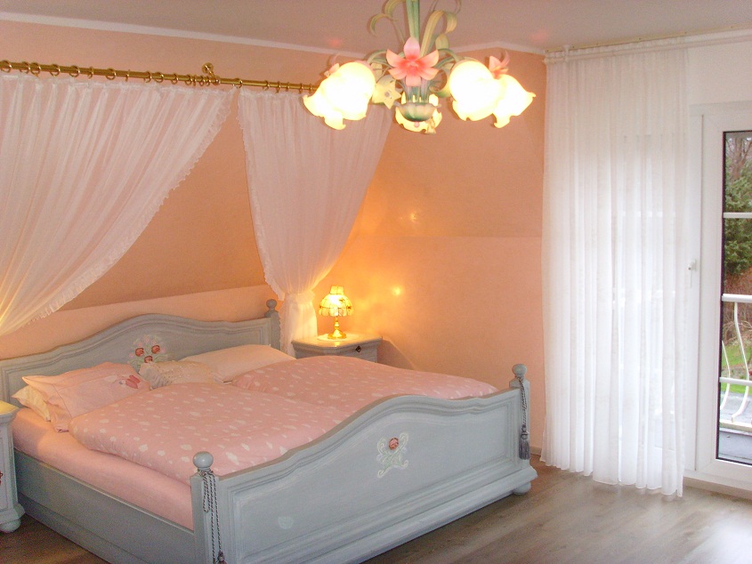 Jasna sypialnia z łóżkiem w stylu vintage, baldachimem z delikatnych firan i kwiecistym żyrandolem.