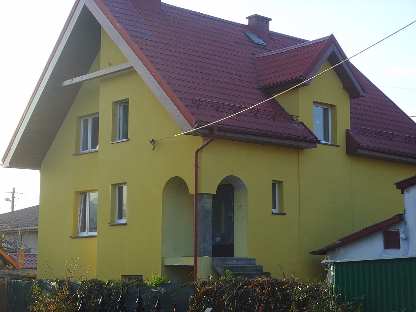 Żółty dom jednorodzinny z czerwoną dachówką, widoczne ocieplenie elewacji, okna PCV, podcień z kolumną i schody wejściowe.