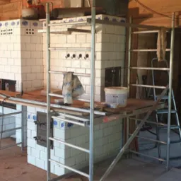 Dwa piece kaflowe w trakcie budowy, białe kafle z niebieskimi akcentami, rusztowania wokół, surowe wnętrze z ceglanymi i drewnianymi ścianami.