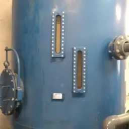 Niebieski zbiornik ciśnieniowy z dwoma wziernikami i przyłączami rurowymi, stojący na trzech nogach w pomieszczeniu technicznym.