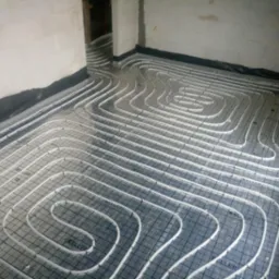 Rury ogrzewania podłogowego ułożone na czarnej folii z siatką w surowym wnętrzu, widoczne białe ściany z bloczków i fragment otworu drzwiowego.