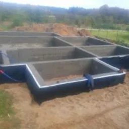 Zalane betonowe fundamenty domu jednorodzinnego z hydroizolacją, widoczne wystające rury instalacyjne, teren budowy z ziemią w tle.