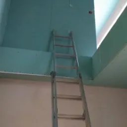 Wnętrze w trakcie prac wykończeniowych: ściany i sufit pokryte płytami gipsowo-kartonowymi, widoczna aluminiowa drabina oparta o ścianę, otwór okienny w suficie.