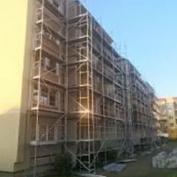 Blok mieszkalny w trakcie prac elewacyjnych, otoczony rusztowaniami, widoczne materiały budowlane na trawniku.