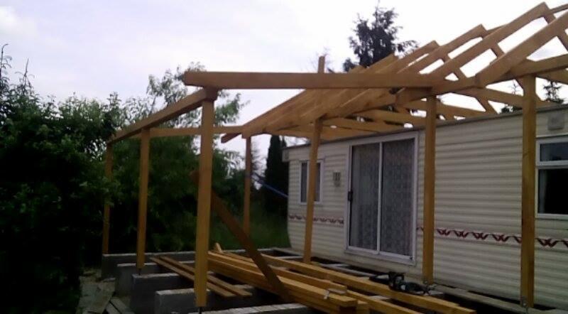 Konstrukcja drewniana zadaszenia w trakcie budowy przy budynku typu barakowóz, widoczne deski i słupy, w tle zieleń i pochmurne niebo.