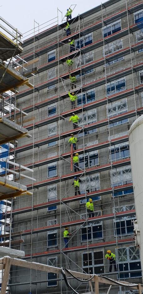 Wielopiętrowe rusztowanie obejmuje budynek w trakcie prac elewacyjnych, z pracownikami w jaskrawych kamizelkach, częściowo zakrytymi oknami i widocznym śniegiem.