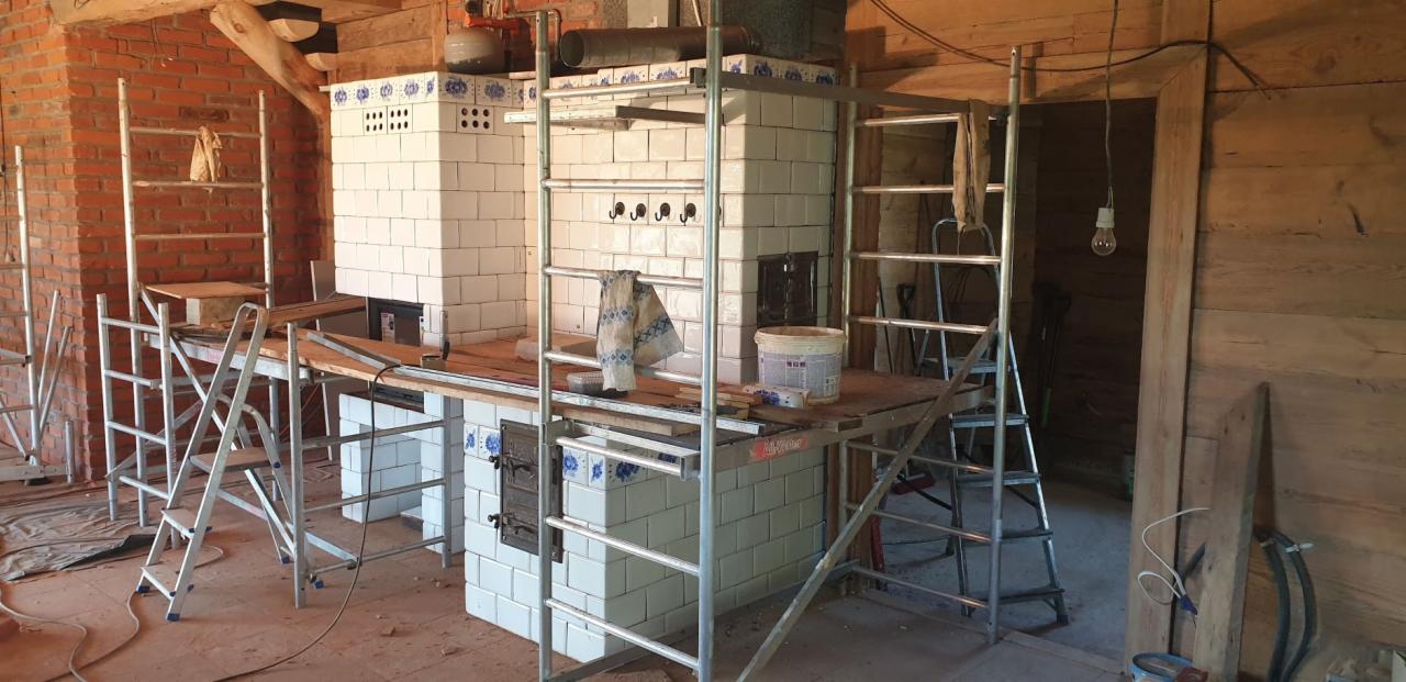 Dwa piece kaflowe w trakcie budowy, białe kafle z niebieskimi akcentami, rusztowania wokół, surowe wnętrze z ceglanymi i drewnianymi ścianami.