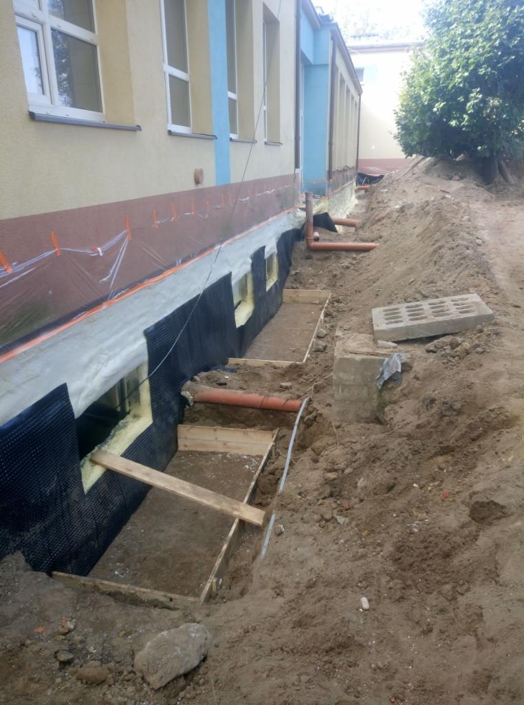 Fundament budynku w trakcie izolacji: widoczne wykopy, folia ochronna, styropian, membrana kubełkowa oraz rury kanalizacyjne.