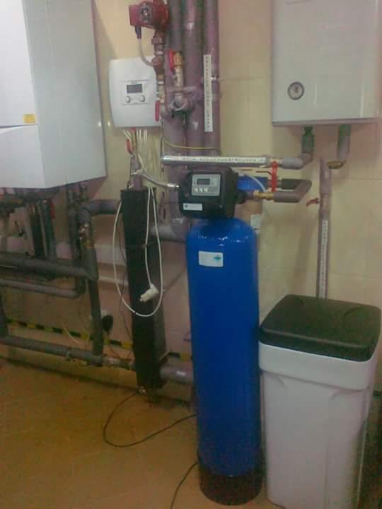 Instalacja uzdatniania wody z niebieskim filtrem i zbiornikiem na sól w kotłowni w Rumi, widoczne rury, zawory i bojlery.
