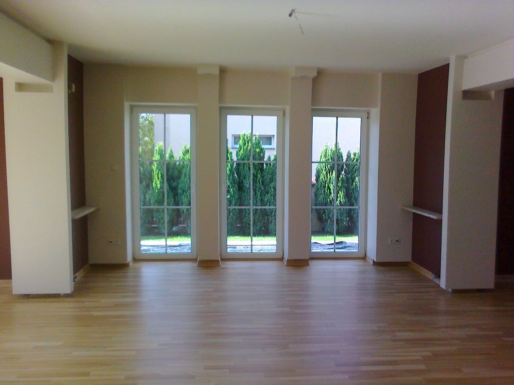 Pusty, jasny pokój z parkietem i trzema dużymi oknami z widokiem na zielone tuje. Ściany w kolorze beżowym i bordowym, z małymi półkami. Widoczne ślady po instalacji oświetlenia na suficie.