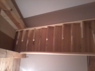 Surowa konstrukcja drewniana antresoli w trakcie budowy, widoczne pionowe i poziome elementy szkieletu, oświetlenie budowlane w tle.