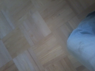 Niejednolicie wycyklinowana, jasna parkietowa podłoga w jodełkę, z widocznym fragmentem spodni osoby wykonującej prace.