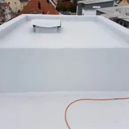 Biały, płaski dach pokryty membraną, z widocznym kominkiem wentylacyjnym i pomarańczowym przewodem na powierzchni, w tle zabudowa miejska.