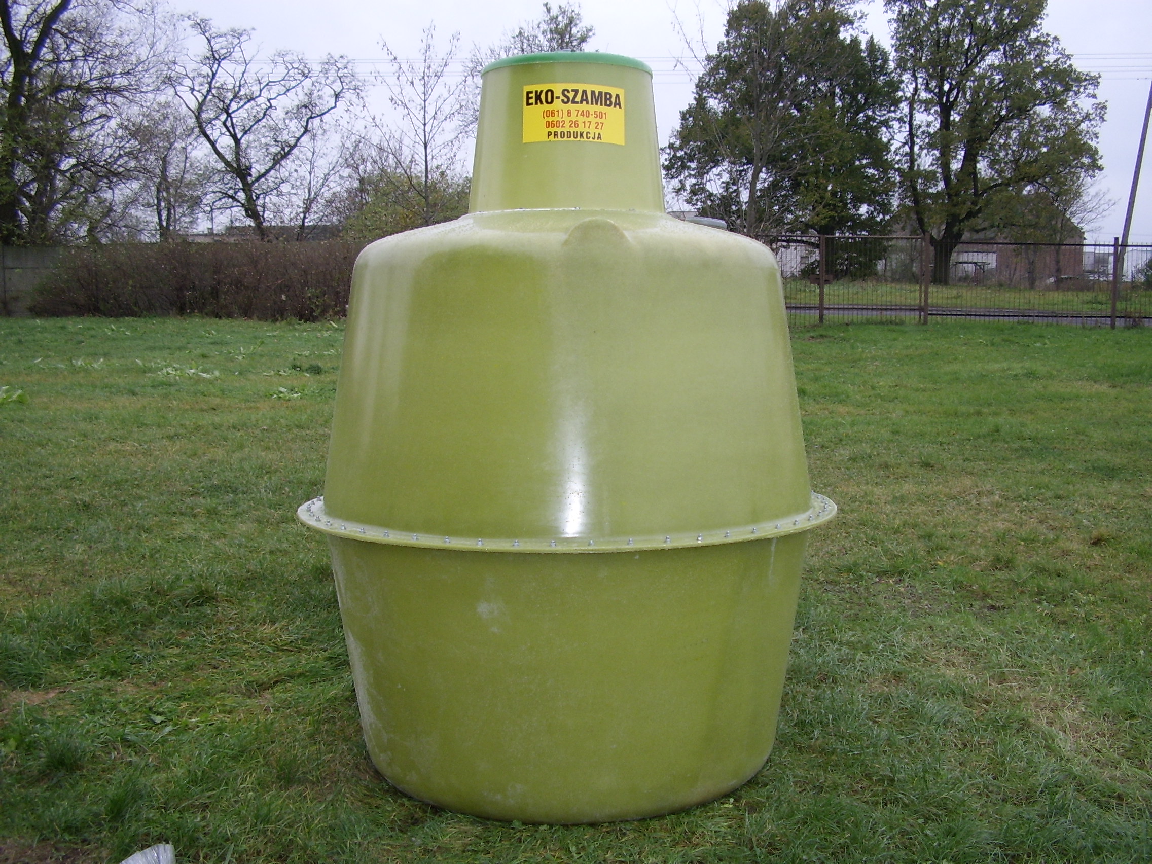 Zielony zbiornik na szambo z naklejką informacyjną, umieszczony na trawniku, z widocznym ogrodzeniem w tle.