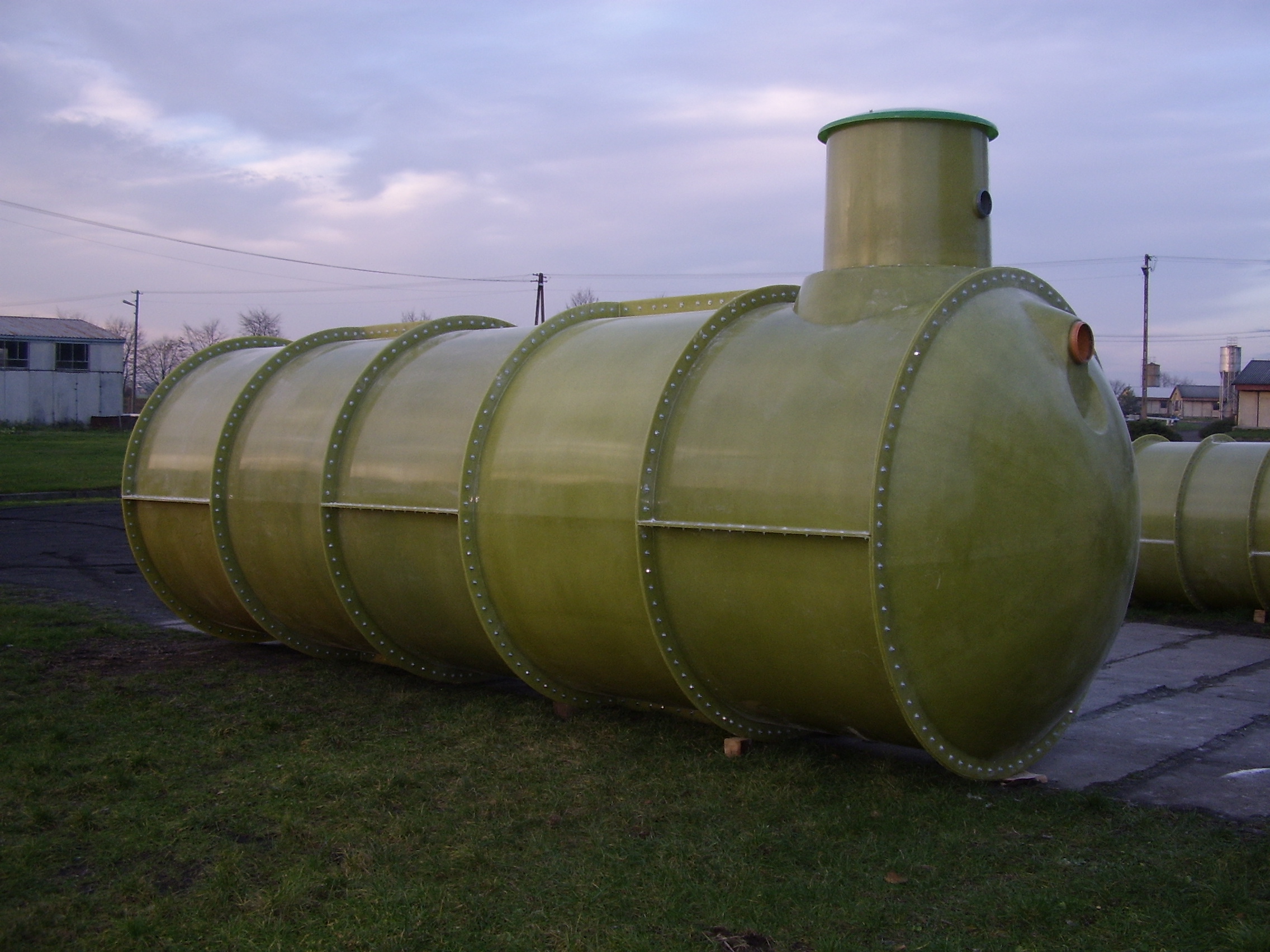 Zielony, cylindryczny zbiornik z włókna szklanego z wystającą rurą, przeznaczony prawdopodobnie na szambo, leżący na trawie przed budynkami.