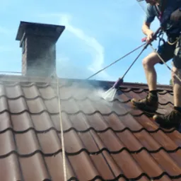 Pracownik w uprzęży, z bliska myje ciemnobrązową dachówkę wodą pod ciśnieniem, obok widoczny komin i fragment błękitnego nieba.