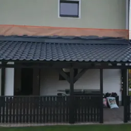 Zadaszenie tarasu z czarną dachówką karpiówką, przylegające do budynku z częściowo zabezpieczoną ścianą, widok z zewnątrz na drewnianą konstrukcję i ogrodzenie.