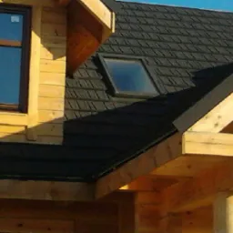 Fragment drewnianego domu z czarnym dachem pokrytym dach&oacute;wką bitumiczną oraz oknem dachowym, widoczne r&oacute;wnież okno w ścianie szczytowej.