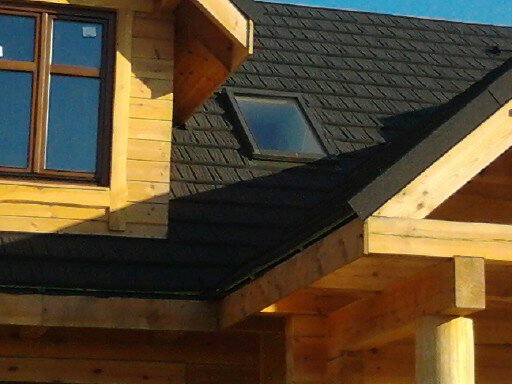 Fragment drewnianego domu z czarnym dachem pokrytym dachówką bitumiczną oraz oknem dachowym, widoczne również okno w ścianie szczytowej.