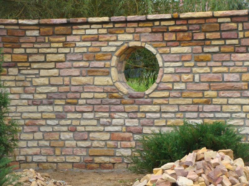 Kamienny mur z okrągłym oknem, wykonany z różnokolorowych kamieni. U dołu roślinność i stos kamieni. Nietypowe wykończenie ogrodu.
