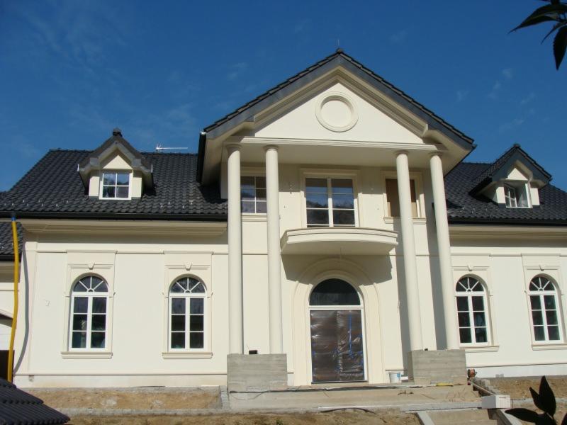 Elewacja domu z kolumnami i balkonem, widoczne okna z łukami, dach z dachówki w kolorze grafitowym. Budynek w stylu klasycystycznym, w jasnej tonacji.