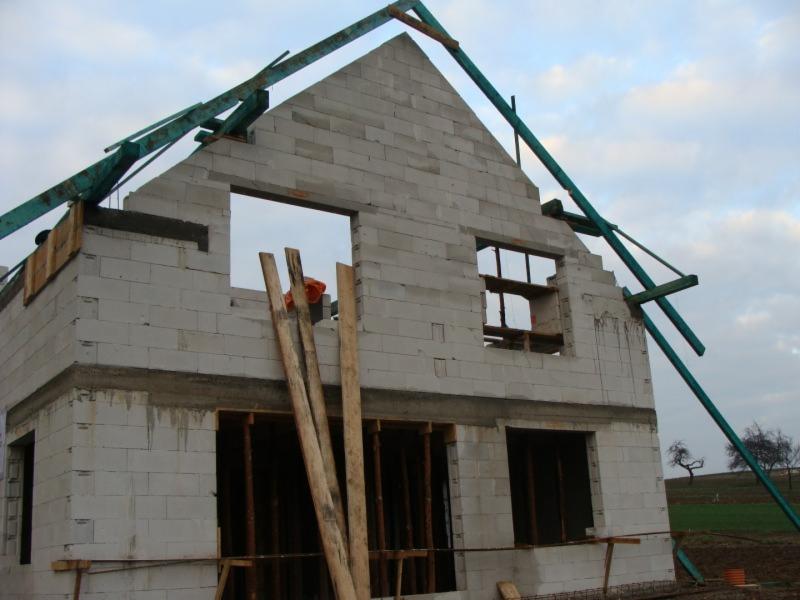 Wznoszenie ścian domu z bloczków betonu komórkowego. Widoczne drewniane stemple i szalunki okienne. W tle pole i drzewo.