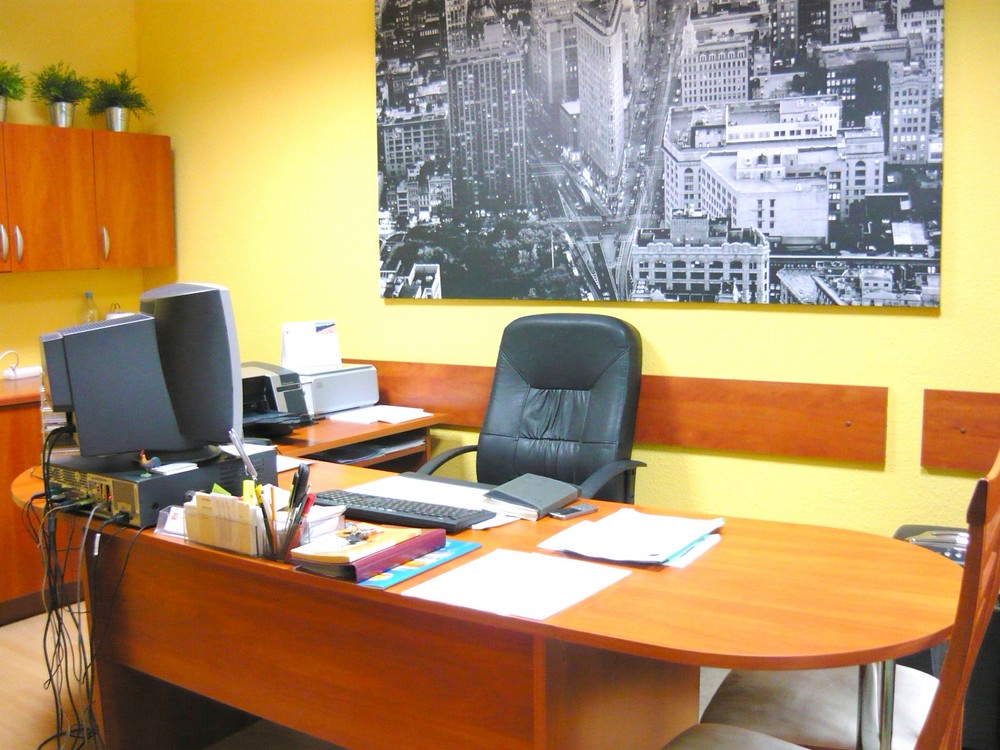 Biuro z żółtymi ścianami, drewnianym biurkiem, czarnym krzesłem biurowym i czarno-białym zdjęciem panoramy miasta na ścianie.