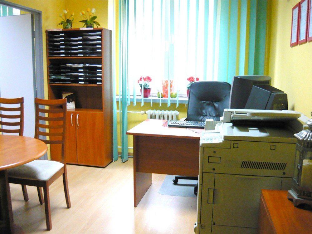 Biuro pośrednictwa nieruchomości w Katowicach: widok na biurko z komputerem, dużą drukarką i szafę z segregatorami w tle, jasne ściany i okno z zasłonami.