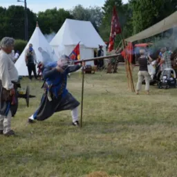 Mężczyzna w stroju historycznym strzela z muszkietu podczas rekonstrukcji bitwy na trawiastym terenie, w tle białe namioty i widzowie.