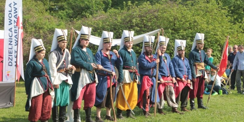 Grupa rekonstruktorów historycznych w strojach z epoki, ustawiona w rzędzie na zielonej trawie, z flagą Dzielnicy Wola w tle, podczas wydarzenia plenerowego.