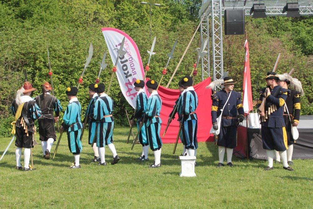 Grupa mężczyzn w historycznych strojach, trzymających halabardy, na tle zieleni i sceny z nagłośnieniem.