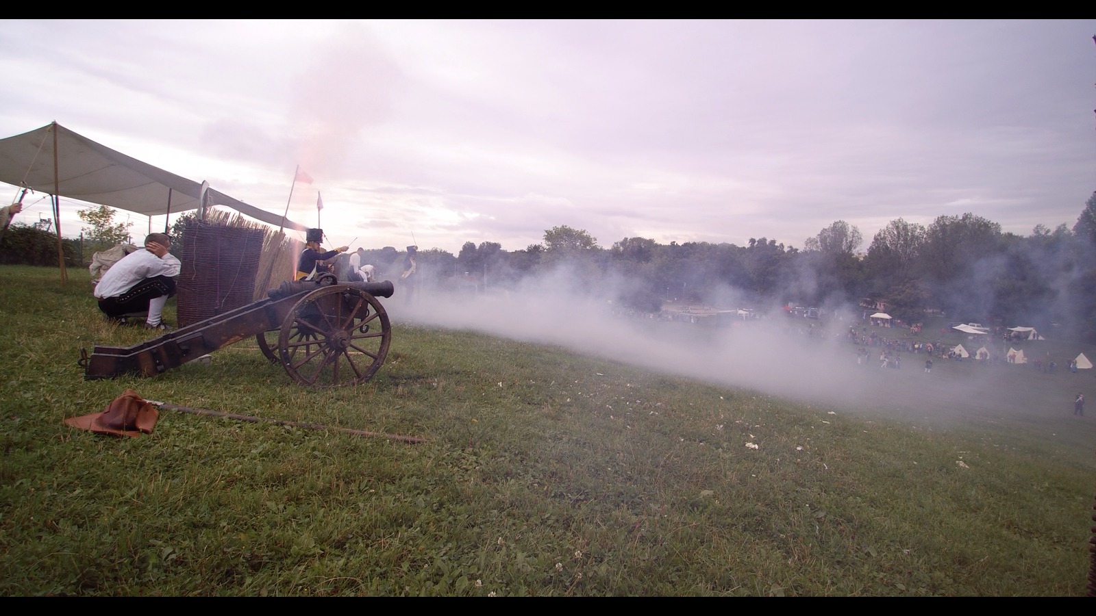 Rekonstrukcja historyczna bitwy: dym z wystrzału armatniego, żołnierze w strojach z epoki, widzowie w tle, na pierwszym planie armata i mężczyzna w stroju ludowym z zatkanymi uszami.